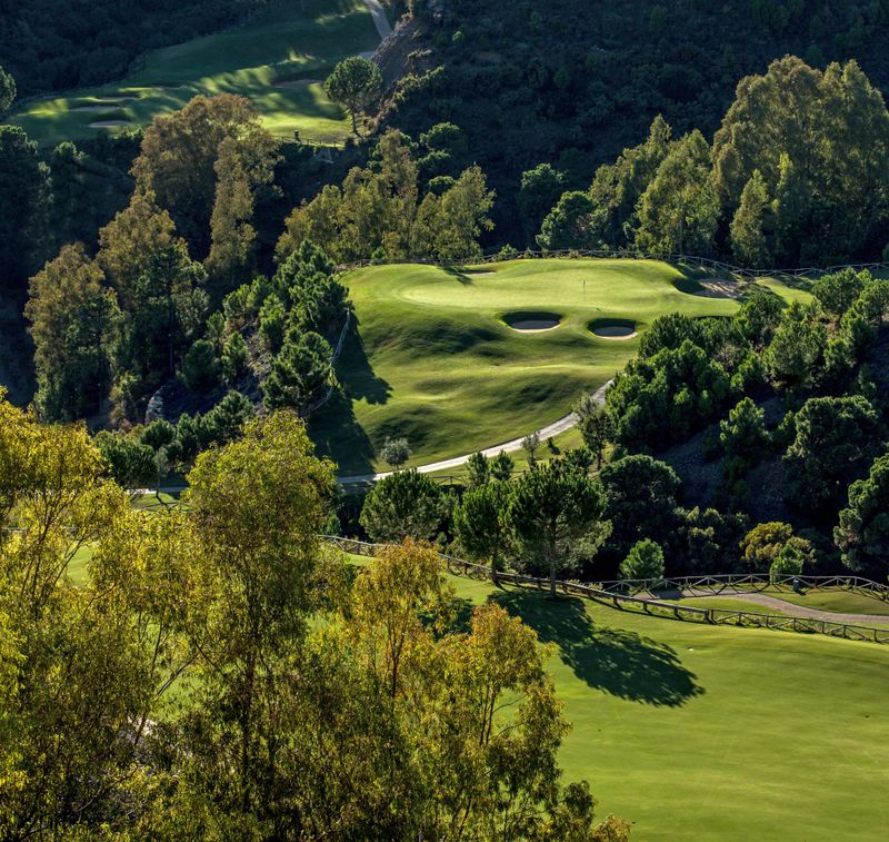 Image of La Zagaleta Golf course, specifically a hilly fairway surrounded by thick green trees and two round bunkers in the middle. A path leads up to the green.